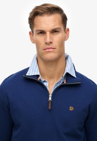 Jersey de zip color navy con un acabado texturizado, que presenta un logo dorado y está superpuesto a una camisa de cuello a rayas en azul claro.
