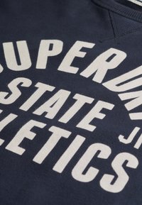 Felpa blu navy con testo bianco in grassetto che recita "SUPERDRY STATE ATHLETICS." Presenta un collo rotondo e un tessuto morbido e testurizzato.