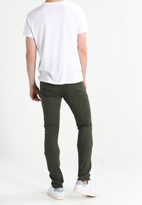 Groene slim-fit broek met achterzakken, gedragen met een simpel wit t-shirt en witte sneakers met lichtblauwe accenten.