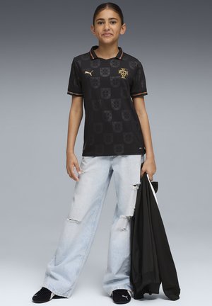 Musta jalkapallopaita, jossa on korostettu kuvio ja kultainen logo, yhdistetty vaaleansinisiin kuluneisiin farkkuihin ja mustiin kenkiin, kädessä musta takki.