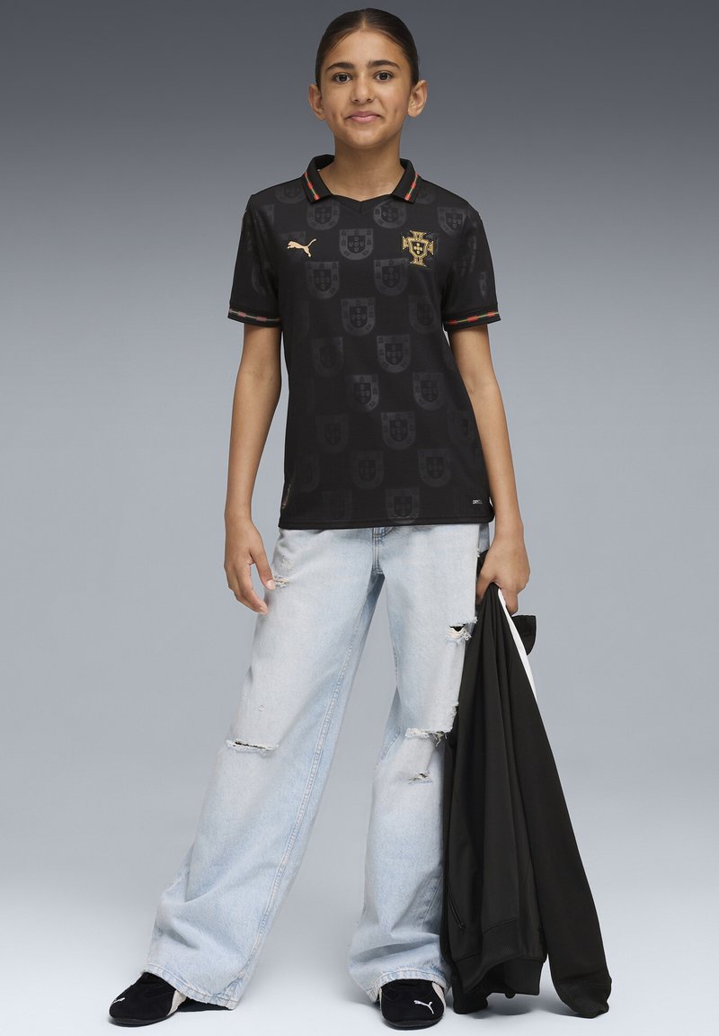 Črna nogometna majica z teksturiranim vzorcem in zlatim logotipom, kombinirana s svetlo modrimi raztrganimi kavbojkami in črnimi čevlji, v roki drži črno jakno.