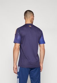 Maglietta sportiva viola con maniche a strisce blu, caratterizzata da un piccolo logo "LA" arancione sul retro del collo. Tessuto liscio e leggero.
