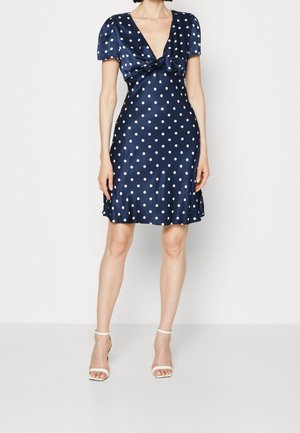 Mujer con un vestido de satén azul marino con lunares blancos, mangas cortas abullonadas, detalle de lazo en la parte delantera y zapatos de tacón blanco con pulsera en el tobillo.