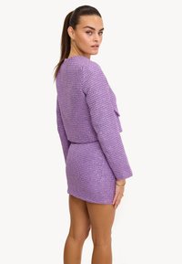 Ensemble tricoté violet en deux pièces comprenant une veste courte et une jupe ajustée. La texture est plissée avec un motif subtil. Accents en matériel doré.
