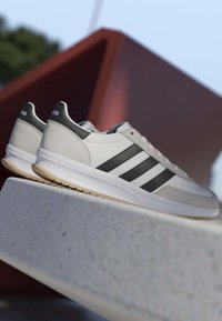 Białe i szare buty sportowe Adidas z czarnymi paskami wystawione na betonowej powierzchni na tle rozmytego, zewnętrznego tła.
