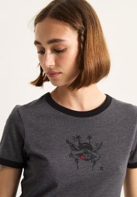 Grå bomulds t-shirt med sort ribbet halsudskæring. Har et grafisk tryk af et kranium med vinger og stjerner i sort og rød på forsiden.
