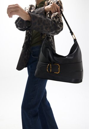 Persona con blazer de estampado de leopardo y vaqueros sostiene un bolso de cuero negro con hebilla dorada sobre fondo blanco.