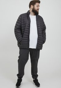 Polstret svart jakke med glidelås, med høy krage og vertikal quiltet design, parret med mørke bukser og svarte joggesko.