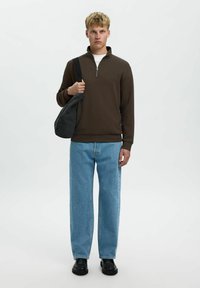 Hnědá svetr na zip, světle modré volné džíny a černé boty. Drží tmavě šedou kabelku přes rameno. Neutrální pozadí.