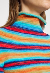 Pull à col roulé multicolore avec des rayures horizontales rouges, bleues, oranges et jaunes. Le tissu semble doux et texturé.