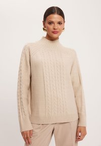 Beżowy sweter w sploty z wysokim kołnierzem, z prążkowanymi mankietami i dolnym wykończeniem. Miękka faktura, luźny fason oraz wyrazisty wzór splotu.