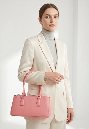 Femme en costume beige et col roulé gris tenant un sac à main rose structuré avec double poignées sur fond blanc.