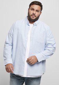 Világoskék, gombos ing, amely texturált anyagból készült, mellzsebével és hosszú ujjú, egy fehér póló fölé rétegezve.