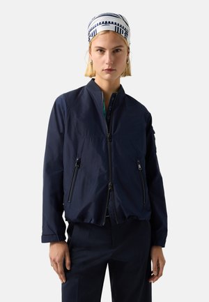 Junge Frau, die eine marineblaue Jacke mit Reißverschluss, passende Hose, silberne Creolen und ein weißes Halstuch mit marineblauen Streifen auf dem Kopf trägt.