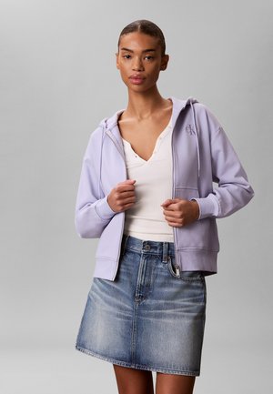 Kobieta nosząca jasnofioletową bluzę z kapturem i zamkiem na białym prążkowanym topie oraz wyblakłą jeansową spódnicę do kolan, stojąca na jednolitym szarym tle.