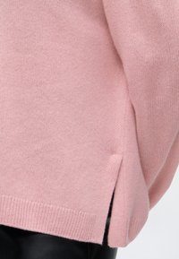 Gros plan sur l'ourlet d'un pull en maille rose douce avec une petite fente latérale porté sur un pantalon noir.