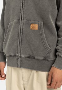 Sudadera con capucha gris, con cremallera frontal, puños acanalados y un parche de cuero beige en el bolsillo. Material suave con un acabado texturizado.