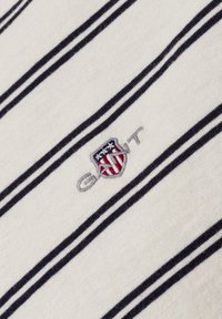 Wit stof met diagonale zwarte strepen en een klein geborduurd schildlogo in rood, wit en blauw met het woord "GANT."