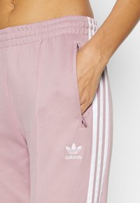 Ljusrosa sweatpants med sidizipar, elastisk midja, tre vita ränder längs sidorna och en liten vit Adidas-logga.