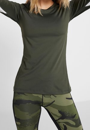 Femme portant un maillot de sport à manches longues vert foncé et des leggings camouflage vert et noir, bras levés sur un fond uni.