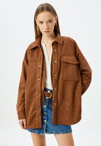 Giacca camicia in finta pelle scamosciata marrone con bottoni a pressione, una tasca sul petto, e una vestibilità rilassata, indossata sopra una maglietta bianca e una gonna di jeans.