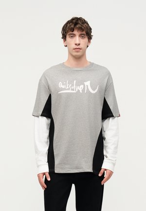 Ung mand med krøllet hår iført lagdelte grå og hvide langærmede trøjer med sorte sidepaneler, stående mod en ensfarvet lys baggrund.