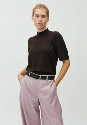 Femme aux cheveux blonds portant un haut noir à manches courtes rentré dans un pantalon large taille haute rose avec une ceinture noire, les mains dans les poches.