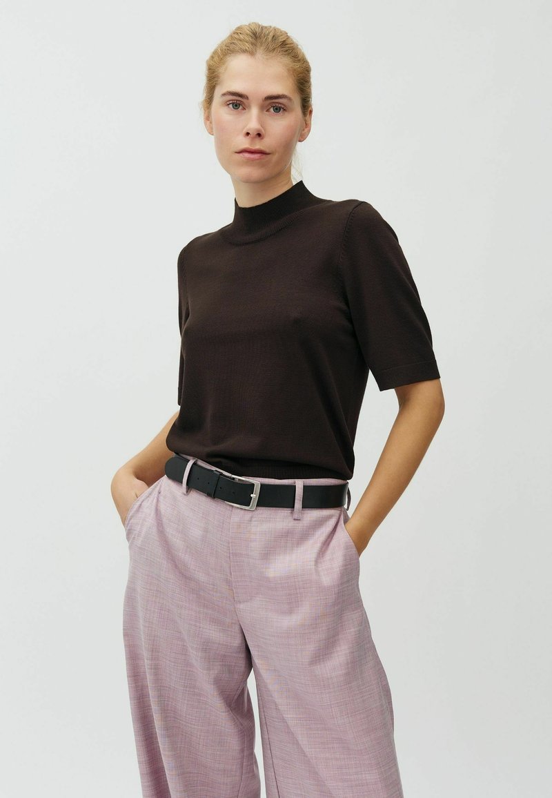 Femme aux cheveux blonds portant un haut noir à manches courtes rentré dans un pantalon large taille haute rose avec une ceinture noire, les mains dans les poches.