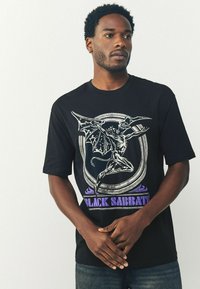 Mand iført en sort Black Sabbath T-shirt med vingerdemon grafik og lilla tekst, stående med hænderne flettet mod en enkel lys baggrund.