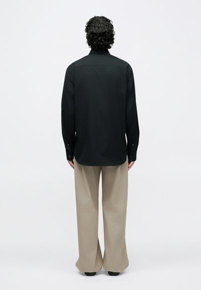Chemise noire à manches longues, coupe droite avec poignets boutonnés, associée à un pantalon beige à jambes larges. Tissu lisse, design minimaliste.