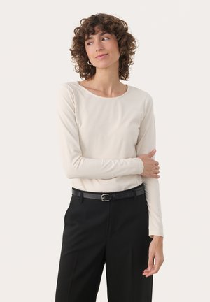 Femme aux cheveux bouclés portant un haut beige à manches longues et un pantalon noir, debout, un bras croisé et regardant sur le côté.