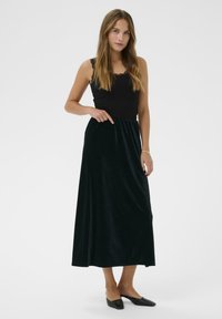 Frau in einem ärmellosen schwarzen Oberteil mit Spitzendetails, einem langen schwarzen Samtrock, schwarzen spitzen Ballerinas und einem goldenen Armband, stehend vor einem weißen Hintergrund.