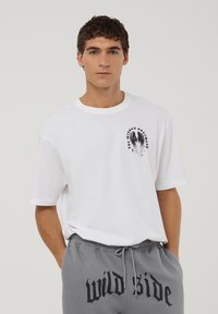 T-shirt bianco in cotone con maniche corte e scollatura rotonda. Presenta un design grafico nero sul lato sinistro del petto. Abbinato a pantaloni della tuta grigi.