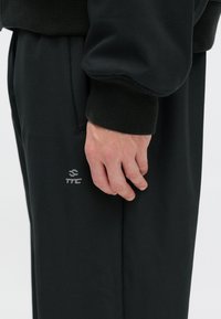 Μαύρες φόρμες με λεία υφή, που διαθέτουν ένα διακριτικό λογότυπο σε γκρι κοντά στην τσέπη και ελαστικά μανίκια. Χέρι που ξεκουράζεται πάνω στο ύφασμα.