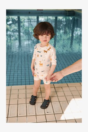 Traje de baño de color claro, de manga corta, con estampados de animales, combinado con zapatos de agua negros. De pie sobre una superficie de baldosas cerca de una piscina.