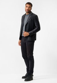 Schwarze Lederblazer mit Reverskragen, einreihiger Knopfschluss und einer Tasche. Getragen über einem grauen Pullover und kombiniert mit schwarzen Hosen und Stiefeln.