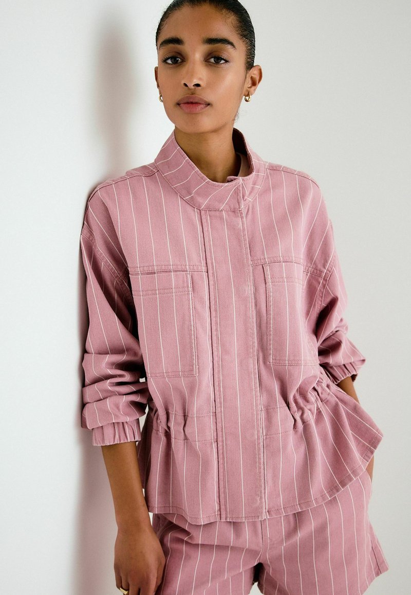 Jeune personne portant une veste rose à rayures fines avec une taille élastique et un short assorti, debout contre un mur blanc les mains dans les poches.