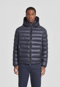 Giubbotto imbottito blu navy con cappuccio, caratterizzato da un design trapuntato, chiusura frontale con zip e tasche laterali con zip. Texture liscia e forma imbottita.