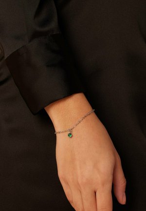 Silberne Kettenarmband mit einem grünen Edelsteinakzent; getragen an einer Hand vor einem dunklen Stoffhintergrund. Glatte Textur und zartes Design.