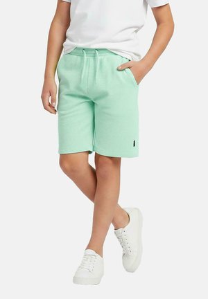 Kind draagt mintgroene shorts met trekkoord, witte sneakers en een wit T-shirt, staat met één hand in de zak.