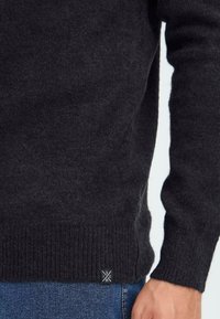 Close-up van een persoon in een zwarte gebreide trui en blauwe jeans, met de mouw, tailleband en een klein zwart label met witte tekst op de trui.