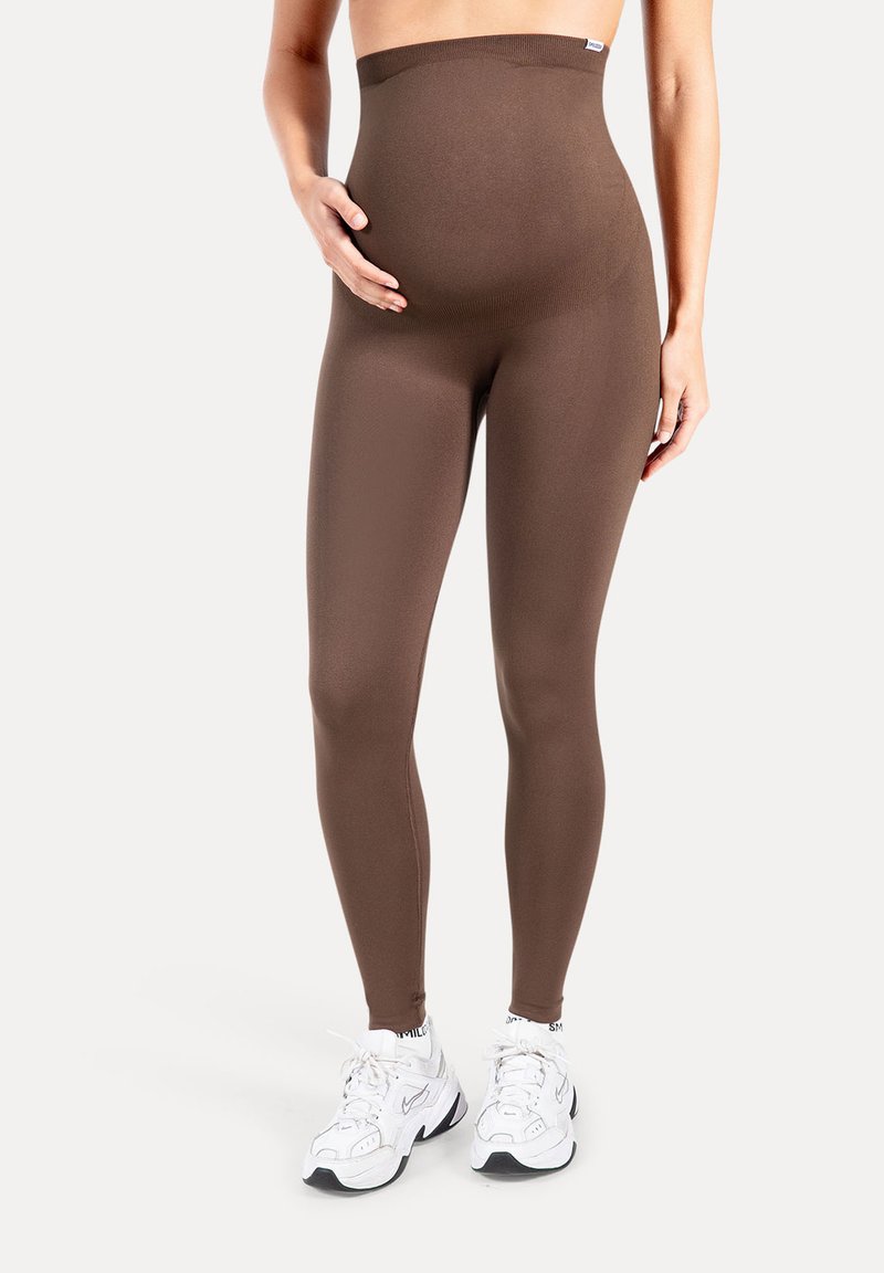 Leggings de maternité marron en tissu lisse et extensible avec une taille haute. Portés avec des chaussures de sport blanches.