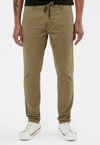 Chinos en coton mélangé khaki avec une taille à cordon, coupe slim et texture subtile, associés à des baskets blanches et une chemise noire.