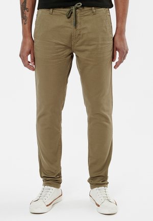 Chinos en coton mélangé khaki avec une taille à cordon, coupe slim et texture subtile, associés à des baskets blanches et une chemise noire.