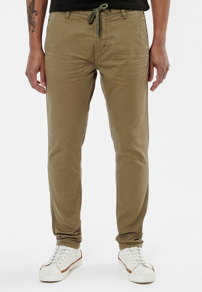 Chinos en coton mélangé khaki avec une taille à cordon, coupe slim et texture subtile, associés à des baskets blanches et une chemise noire.