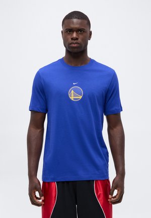 Homme portant un t-shirt bleu avec un logo jaune de pont et un short de sport rouge et noir, debout sur un fond blanc.