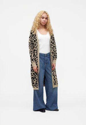 Jeune femme aux longs cheveux blonds bouclés portant un long cardigan imprimé léopard, un top blanc, un jean bleu à jambes larges et des chaussures noires.