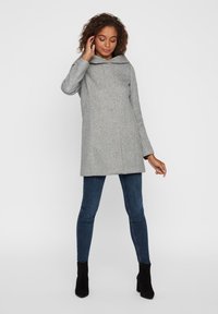 Cappotto grigio in misto lana con cappuccio, caratterizzato da una linea dritta, tasche laterali e una texture morbida, abbinato a jeans scuri e stivaletti neri alla caviglia.