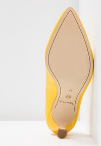 Chaussure à talon haut jaune à bout pointu reposant sur le côté de la semelle contre une surface blanche, montrant la taille 37 et la marque "Tamaris" en dessous.