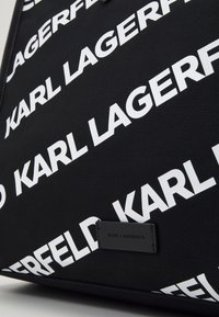 Svart väska med upprepat vitt "KARL LAGERFELD"-logotyp. Tillverkad av strukturerad tyg, med en svart etikett med präglad logotyp längst ner.